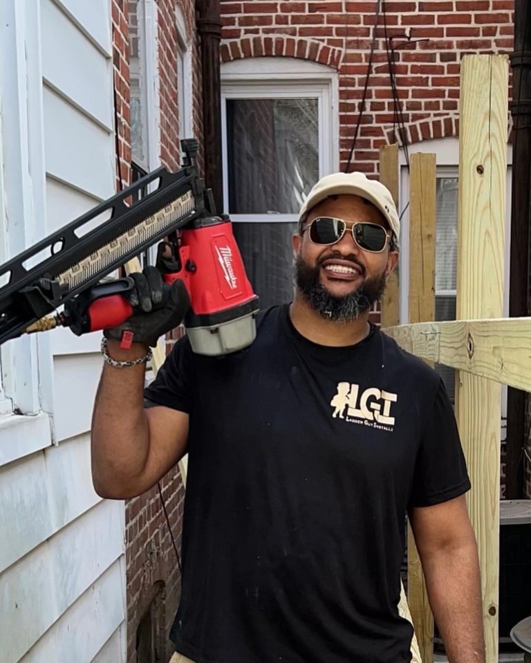 Man in black #LGT shirt and cap holding red power tool on shoulder, standing in front of brick house with white trim