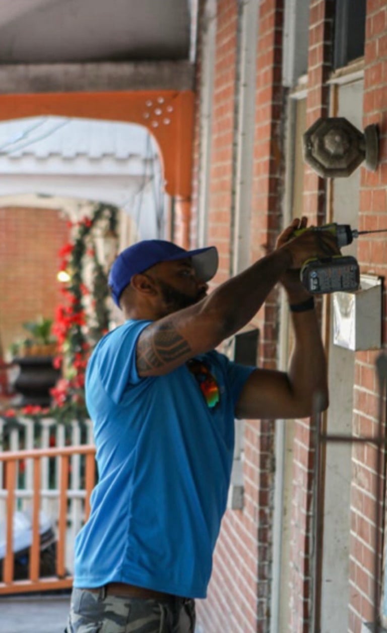 Delivery person in blue shirt and cap scanning package at brick doorway with holiday wreath in background